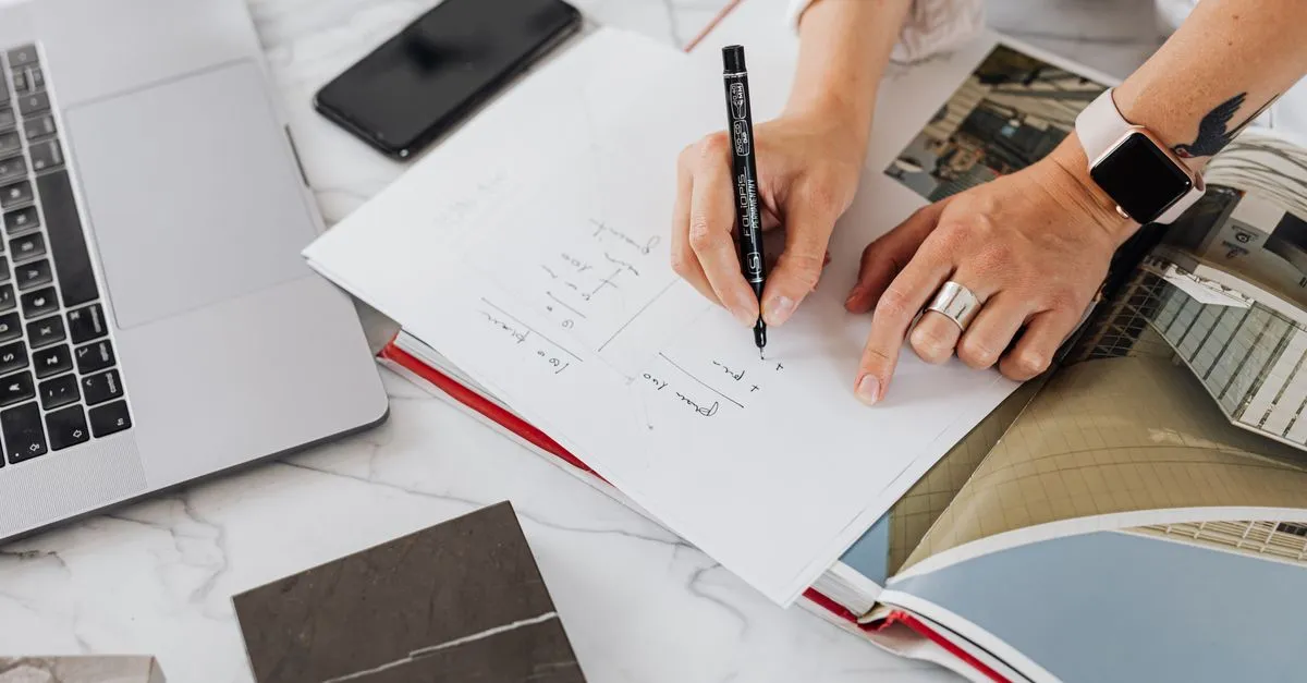 Hands writing on paper at a desk with laptop and smartphone in a modern office setting.