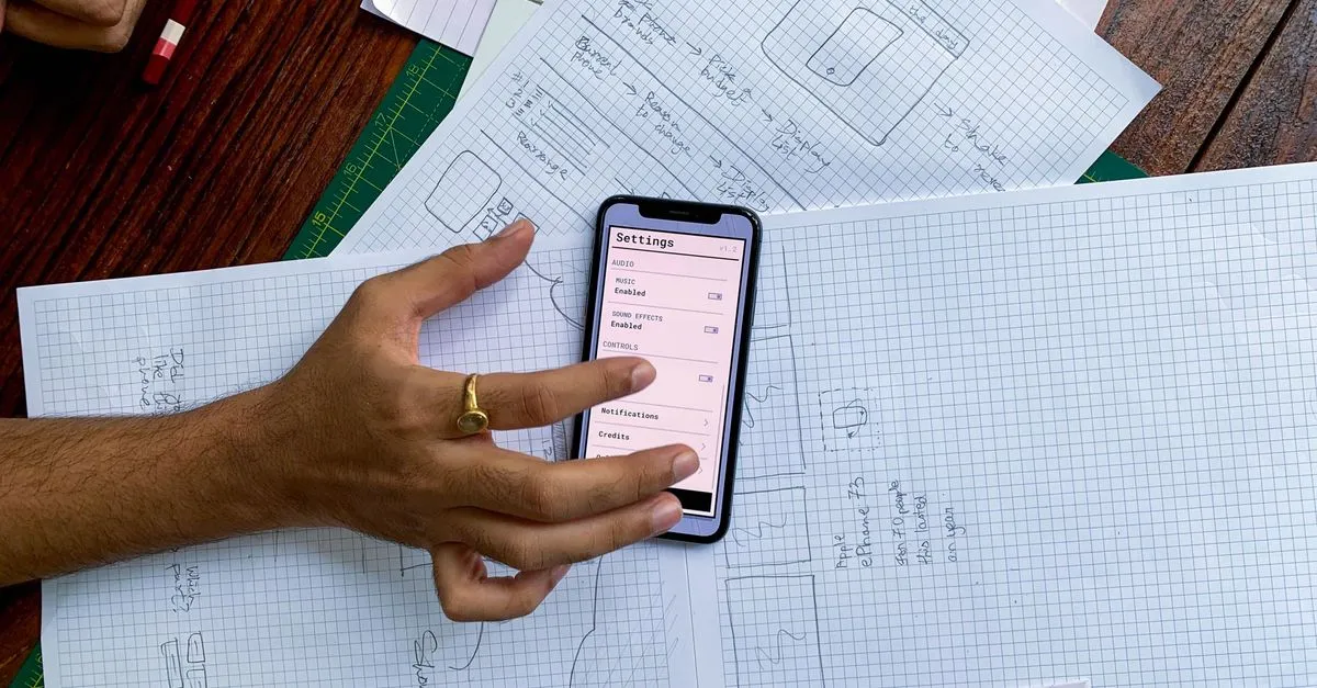 Hand holding smartphone over app design sketches on papers, top view.